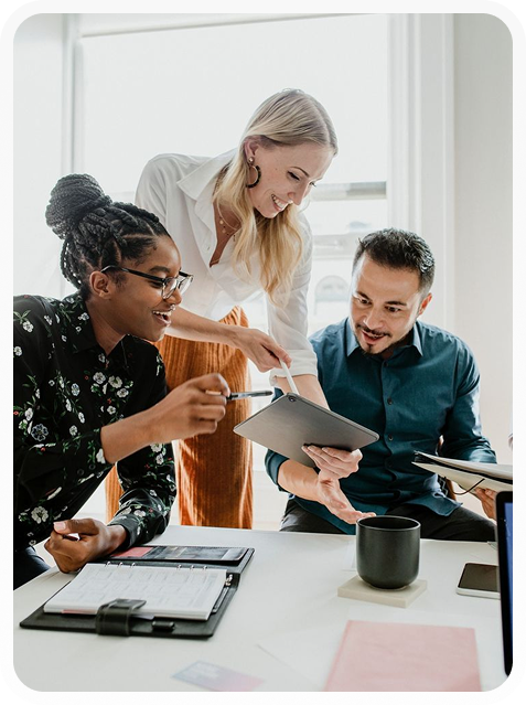 Three diverse coworkers discussing and smiling while reviewing information on a tablet in a bright office.
